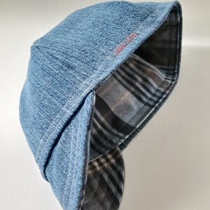 May include: A blue denim flat cap featuring a structured crown and a curved brim. The interior lining displays a grey, black, and white plaid pattern. The text "CORAL MOON" is embroidered in small red letters on the side of the cap's brim.