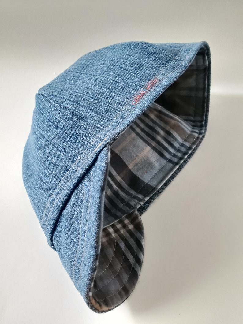 May include: A blue denim flat cap featuring a structured crown and a curved brim. The interior lining displays a grey, black, and white plaid pattern. The text "CORAL MOON" is embroidered in small red letters on the side of the cap's brim.
