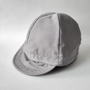 May include: A light gray cycling cap featuring a short, rounded brim and a multi-paneled crown. The fabric displays decorative zigzag stitching along the brim and the lower edge of the crown, with small red text embroidered on the side panel.