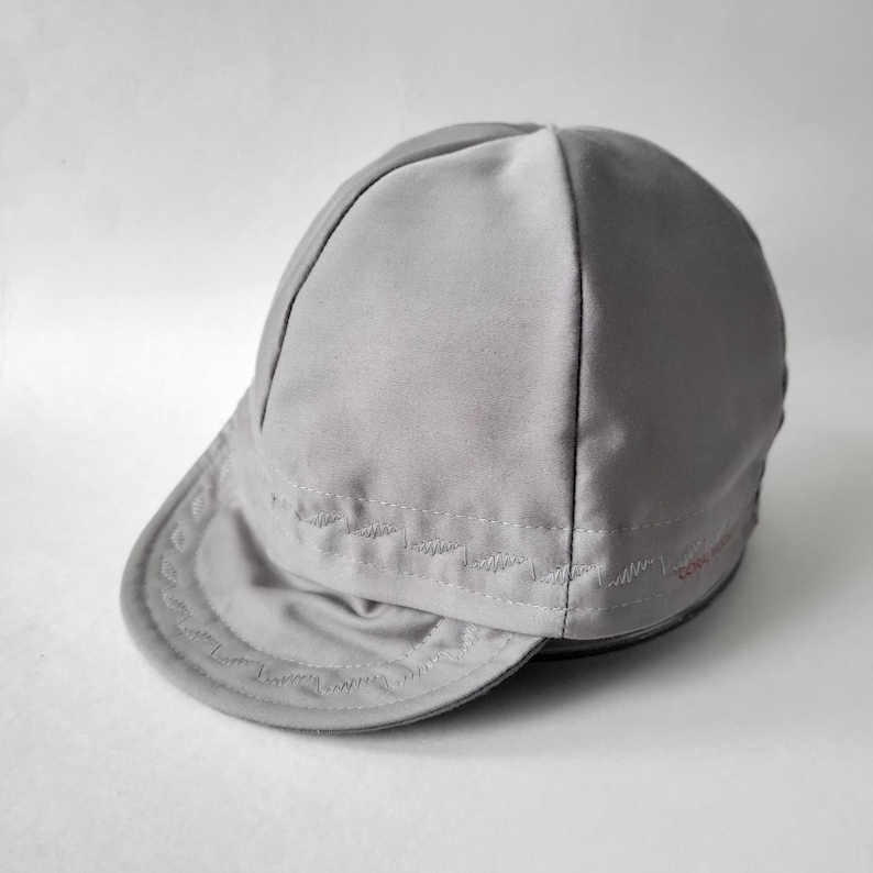 May include: A light gray cycling cap featuring a short, rounded brim and a multi-paneled crown. The fabric displays decorative zigzag stitching along the brim and the lower edge of the crown, with small red text embroidered on the side panel.