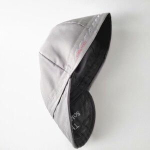 May include: A light gray flat cap with a smooth, solid-colored exterior. The interior lining is dark gray and features visible stitching along the seams. The text "THE SOM" is printed in white on the inner brim, with red embroidery detail.