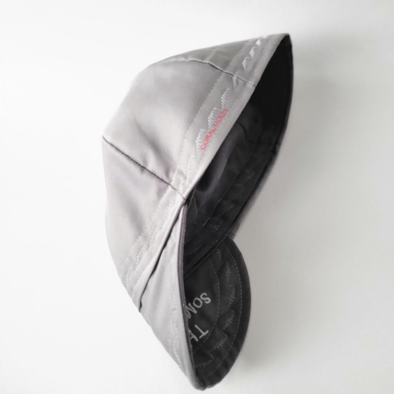 May include: A light gray flat cap with a smooth, solid-colored exterior. The interior lining is dark gray and features visible stitching along the seams. The text "THE SOM" is printed in white on the inner brim, with red embroidery detail.