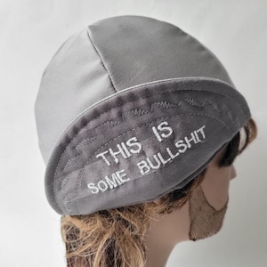 May include: A light gray skull cap featuring a folded brim with the embroidered white text "THIS IS SOME BULLSHIT" across the front. The cap is constructed from a smooth fabric with visible stitching along the brim and crown seams.