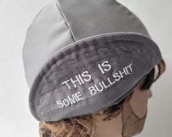 May include: A light gray skull cap featuring a folded brim with the embroidered white text "THIS IS SOME BULLSHIT" across the front. The cap is constructed from a smooth fabric with visible stitching along the brim and crown seams.