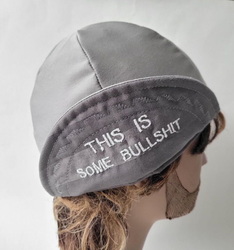 May include: A light gray skull cap featuring a folded brim with the embroidered white text "THIS IS SOME BULLSHIT" across the front. The cap is constructed from a smooth fabric with visible stitching along the brim and crown seams.