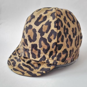 May include: A cap with a leopard print pattern in shades of brown and black. The cap has a short brim and a rounded crown. The fabric appears to be a soft material. The cap is designed to be worn on the head.