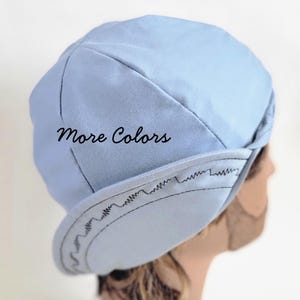 Puede incluir: Una gorra de color azul claro con una corona estructurada y una visera curva con costuras en zigzag negras visibles a lo largo del borde. La tela tiene una textura suave y el texto "More Colors" está impreso en negro en el lateral.