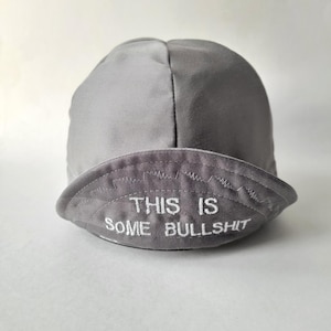 May include: A grey cycling cap with a short, curved brim. The front of the brim features white embroidered text that reads THIS IS SOME BULLSHIT, positioned below a decorative zigzag stitch line that spans the width of the brim.