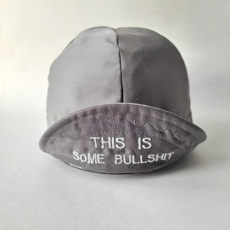 May include: A grey cycling cap with a short, curved brim. The front of the brim features white embroidered text that reads THIS IS SOME BULLSHIT, positioned below a decorative zigzag stitch line that spans the width of the brim.