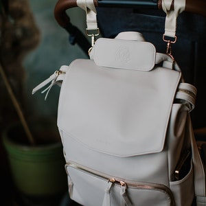 May include: A light beige diaper bag backpack with a rose logo and gold zipper pulls. The bag is attached to a stroller handle with straps.