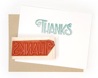 May include: A wooden stamp with the word "THANKS" in a stylized font. The stamp is red and has been used to create an impression on a white card.