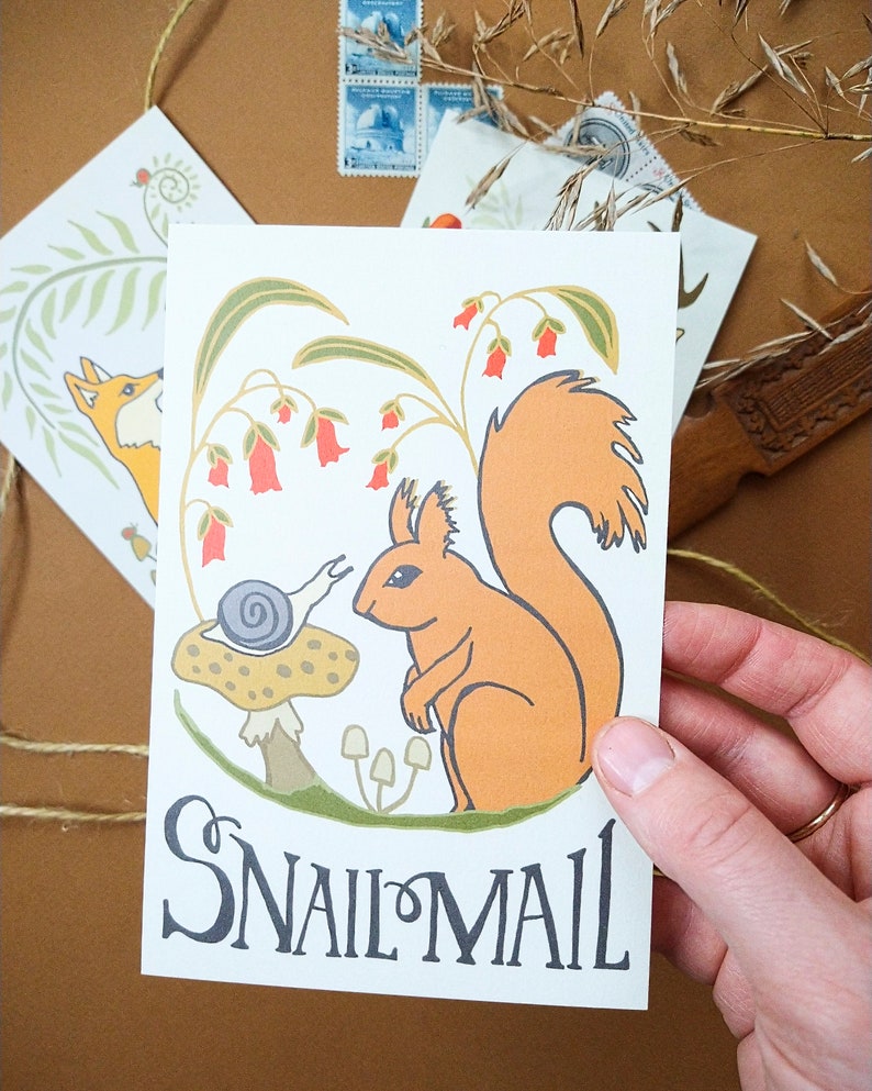 May include: A white card with an illustration of a red squirrel and a snail on a mushroom. The squirrel is sitting on a branch with red flowers. The text "Snail Mail" is written in black at the bottom of the card.