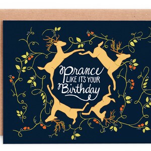 Puede incluir: Una tarjeta de cumpleaños azul marino con un sobre marrón. La tarjeta presenta cuatro ciervos saltando en círculo, con las palabras "Prance like it's your Birthday" en escritura blanca. Acentos florales verdes y naranjas rodean los ciervos.