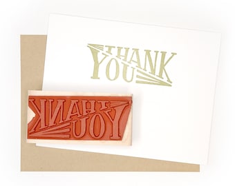 May include: A wooden stamp with the words "THANK YOU" in red lettering. The stamp is on a brown envelope with a white card that says "THANK YOU" in gold lettering.
