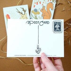 May include: A white postcard with a black and white illustration of mushrooms hanging from a string. The postcard says "POSTCARD" in black cursive writing. There is a black and white stamp that says "PLACE STAMP HERE" in the upper right corner. The postcard is made by Peppercorn Paper.