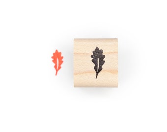 May include: A wooden stamp with a black leaf design. A small, orange leaf impression is next to the stamp. The stamp is made of light-colored wood and is cylindrical in shape. The leaf design is a simple, stylized oak leaf.