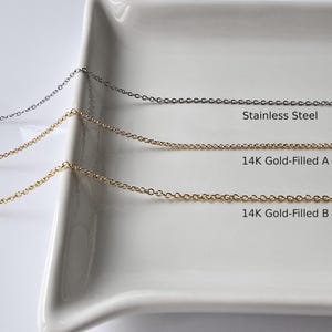 May include: Three chains of different materials and thicknesses on a white surface. The top chain is stainless steel, the middle chain is 14K gold-filled and dainty, and the bottom chain is 14K gold-filled and thick.