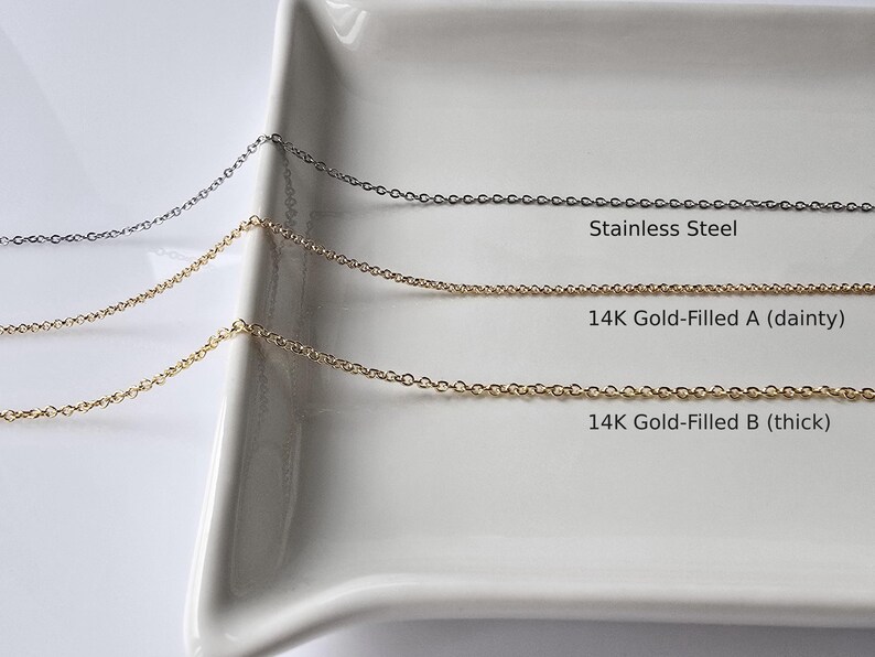 May include: Three chains of different materials and thicknesses on a white surface. The top chain is stainless steel, the middle chain is 14K gold-filled and dainty, and the bottom chain is 14K gold-filled and thick.