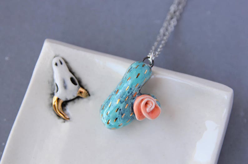 May include: A turquoise ceramic cactus pendant with gold accents and a pink ceramic flower, hanging from a silver chain.