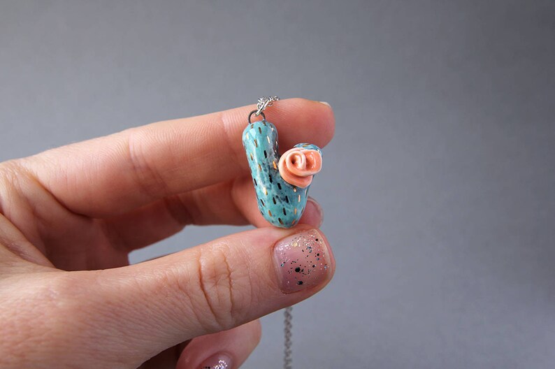 May include: A turquoise and gold cactus pendant necklace with a pink flower on top. The necklace is on a silver chain.
