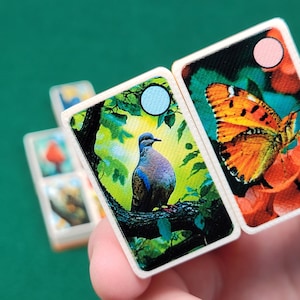 May include: Two dominoes with colorful images. One domino features a blue and gray dove perched on a branch with green leaves. The other domino features an orange butterfly with black markings on a red flower.