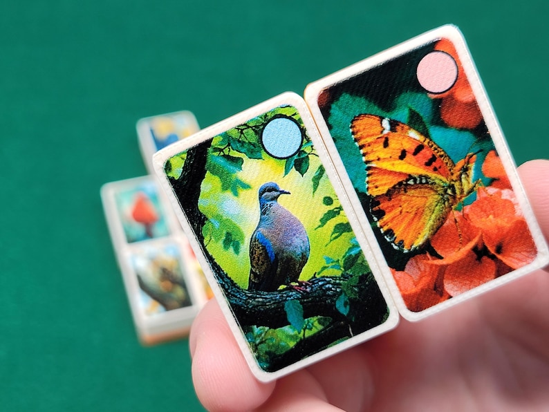 May include: Two dominoes with colorful images. One domino features a blue and gray dove perched on a branch with green leaves. The other domino features an orange butterfly with black markings on a red flower.