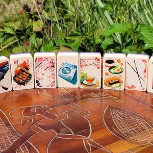 May include: A set of ten dominoes with images of Japanese culture, including a fan, food, cherry blossoms, a teacup, chopsticks, and a traditional Japanese building. The dominoes are white with purple edges.