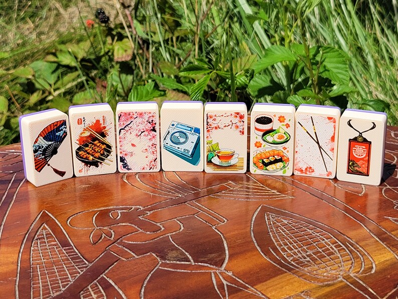 May include: A set of ten dominoes with images of Japanese culture, including a fan, food, cherry blossoms, a teacup, chopsticks, and a traditional Japanese building. The dominoes are white with purple edges.