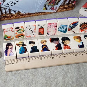 May include: A set of 10 white dominoes with colorful illustrations. The dominoes feature images of food, flowers, a camera, a fan, and anime characters. The dominoes are arranged in a row on a wooden ruler.