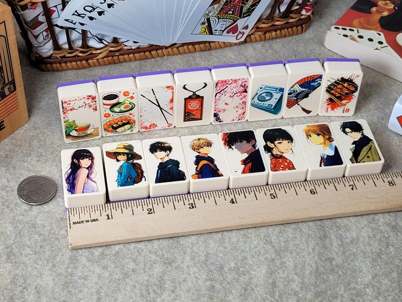 May include: A set of 10 white dominoes with colorful illustrations. The dominoes feature images of food, flowers, a camera, a fan, and anime characters. The dominoes are arranged in a row on a wooden ruler.