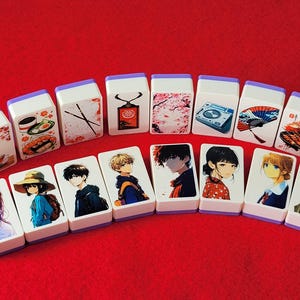 May include: A set of 14 white tiles with purple trim, each featuring a different design. Some tiles have illustrations of anime characters, while others have images of food, flowers, and other objects. The tiles are arranged in a row on a red surface.
