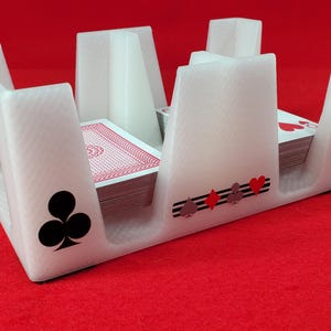 May include: A white plastic playing card holder with four distinct slots, each containing a deck of cards. The exterior features black and red suit symbols including clubs, diamonds, spades, and hearts printed on the front and side panels.
