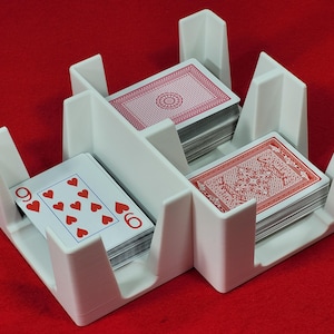 May include: A white plastic card holder with four compartments, each holding a deck of playing cards. The cards have red backs and the face-up cards show the nine of hearts. The holder sits on a red surface.