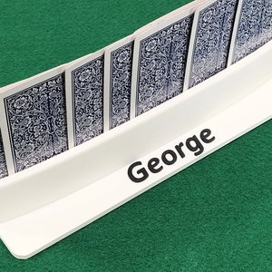 May include: A white card holder with the name "George" printed on the front. The holder is filled with playing cards, all facing down.