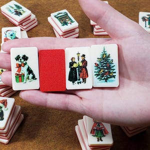May include: A set of Christmas-themed dominoes with images of a dog, a Christmas tree, carolers, and a cat. The dominoes are white with red trim and are held in a hand.