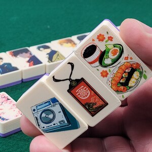 May include: A set of four white dominoes with colorful illustrations. One domino features a camera, another has a necklace with a tag, and the last two have Japanese food illustrations.