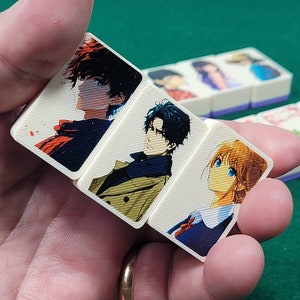 May include: Four white rectangular tiles with colorful anime character illustrations. The tiles are held in a hand. The characters are drawn in a manga style.