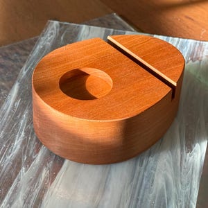 May include: A wooden phone stand in a warm reddish-brown hue. The stand has a rounded shape with a circular cutout and a slot for holding a phone. The wood grain is visible, adding to its natural aesthetic. The stand is designed to hold a phone upright.