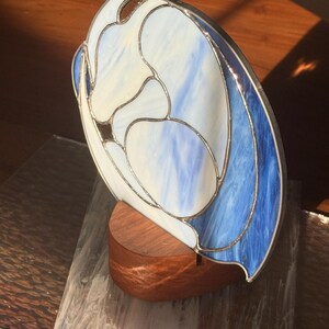 May include: A stained glass sculpture with a flowing design in shades of white and blue, mounted on a wooden base. The glass has a silver metal border. The sculpture is a decorative art piece.