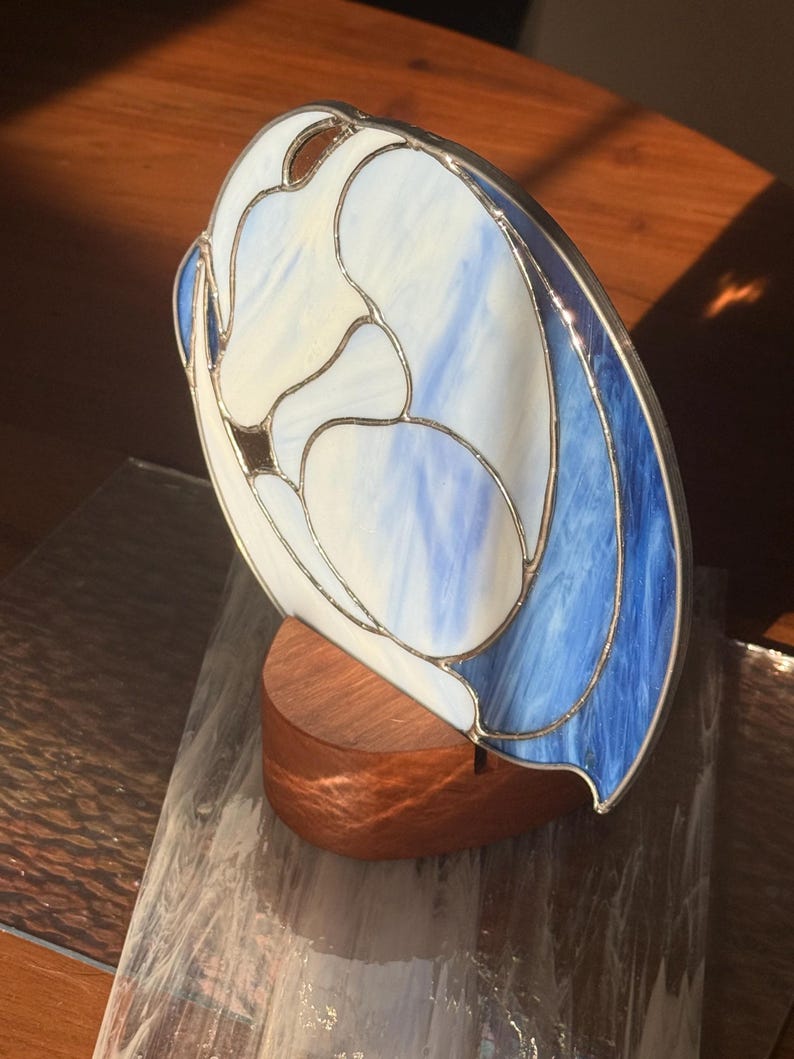 May include: A stained glass sculpture with a flowing design in shades of white and blue, mounted on a wooden base. The glass has a silver metal border. The sculpture is a decorative art piece.