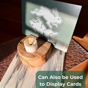 May include: A wooden candle holder with a lit, white candle and a decorative card featuring two white rabbits and the text "Hare + Hog." The card is displayed in the holder. The text "Can Also be Used to Display Cards or Art Prints!" is also visible.
