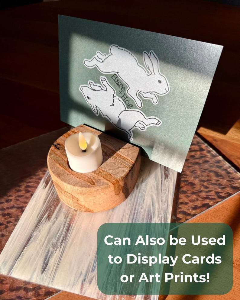 May include: A wooden candle holder with a lit, white candle and a decorative card featuring two white rabbits and the text "Hare + Hog." The card is displayed in the holder. The text "Can Also be Used to Display Cards or Art Prints!" is also visible.