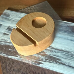 May include: A light-colored wooden phone stand with a circular hole and a slot for holding a phone. The stand is made of a light-colored wood and has a smooth finish. The stand is designed to hold a phone upright.