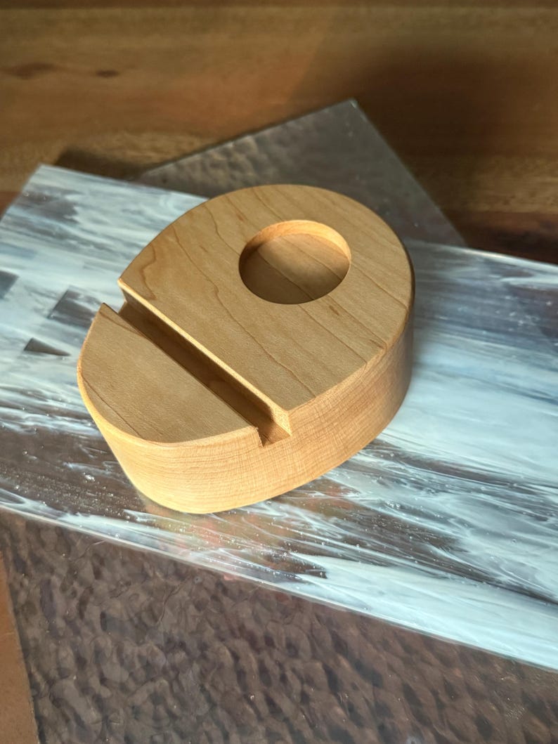 May include: A light-colored wooden phone stand with a circular hole and a slot for holding a phone. The stand is made of a light-colored wood and has a smooth finish. The stand is designed to hold a phone upright.