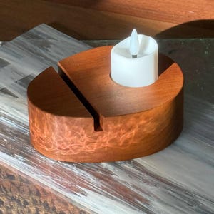 May include: A wooden candle holder with a slot and a white LED candle. The candle holder is made of reddish-brown wood and has a circular design. The LED candle has a flickering flame effect.