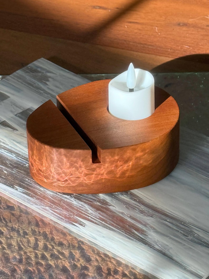 May include: A wooden candle holder with a slot and a white LED candle. The candle holder is made of reddish-brown wood and has a circular design. The LED candle has a flickering flame effect.
