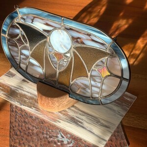 May include: An oval stained glass panel with a blue border, featuring a central white circle and geometric patterns in gold, brown, and white. The panel is displayed on a wooden surface with a wooden support.