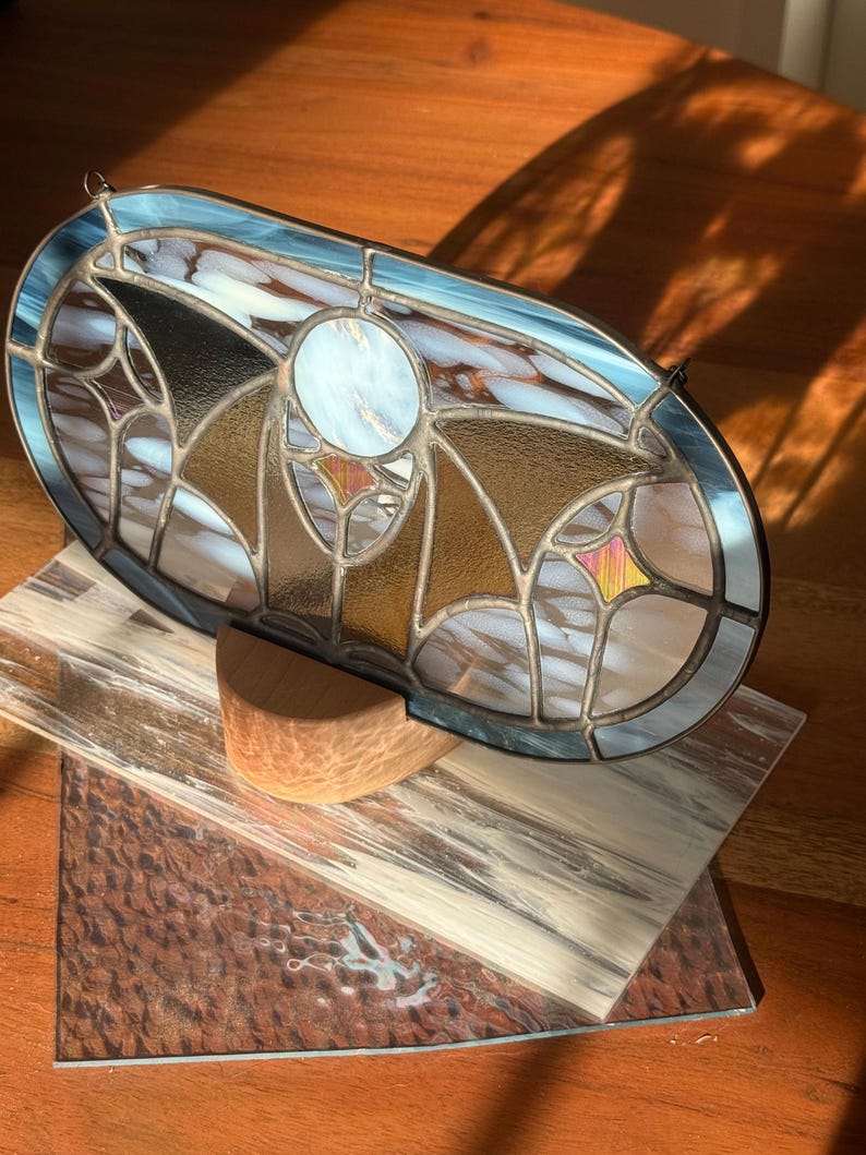May include: An oval stained glass panel with a blue border, featuring a central white circle and geometric patterns in gold, brown, and white. The panel is displayed on a wooden surface with a wooden support.