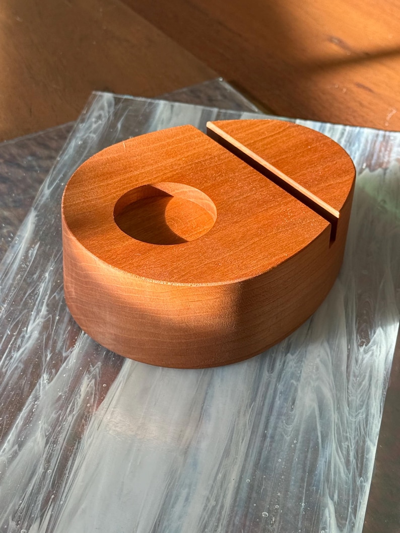 May include: A wooden phone stand in a warm reddish-brown hue. The stand has a rounded shape with a circular cutout and a slot for holding a phone. The wood grain is visible, adding to its natural aesthetic. The stand is designed to hold a phone upright.