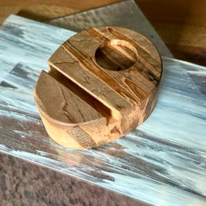 May include: A wooden phone stand with a natural wood grain pattern. The stand has a slot to hold a phone and a circular opening. The wood tones are light and dark brown. The stand is on a patterned surface.